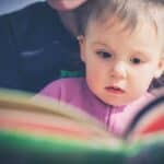 A toddler reading a book sitting on an adult's lap