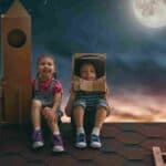 A girl and boy sit on top of a roof with a cardboard spaceship and space helmet