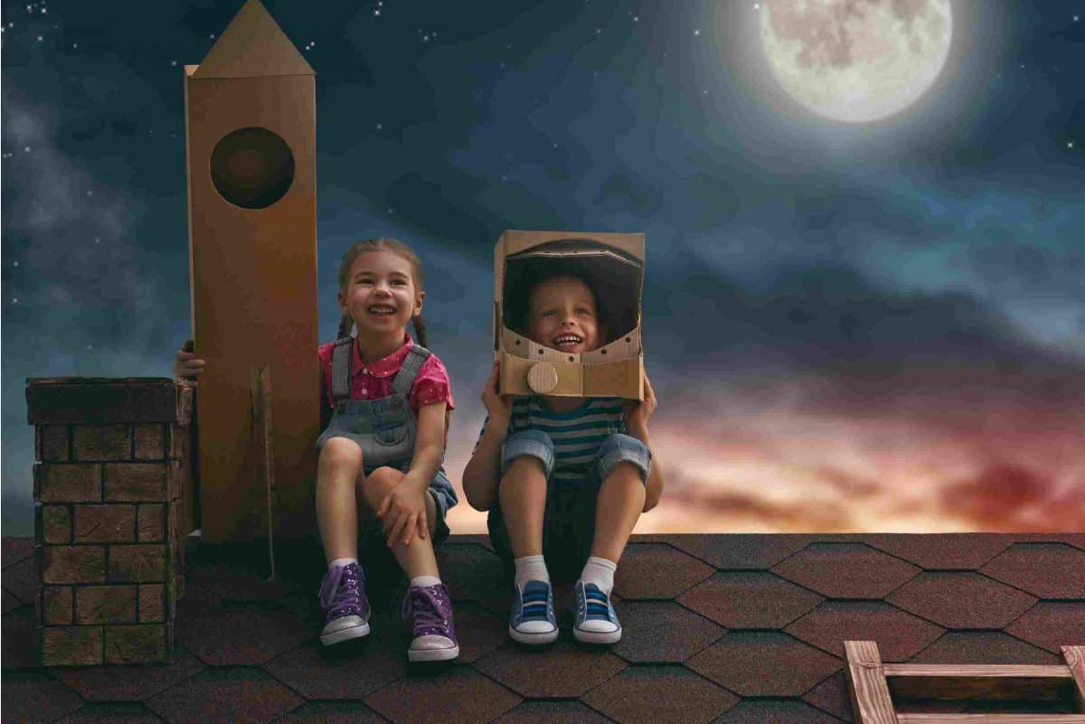 A girl and boy sit on top of a roof with a cardboard spaceship and space helmet